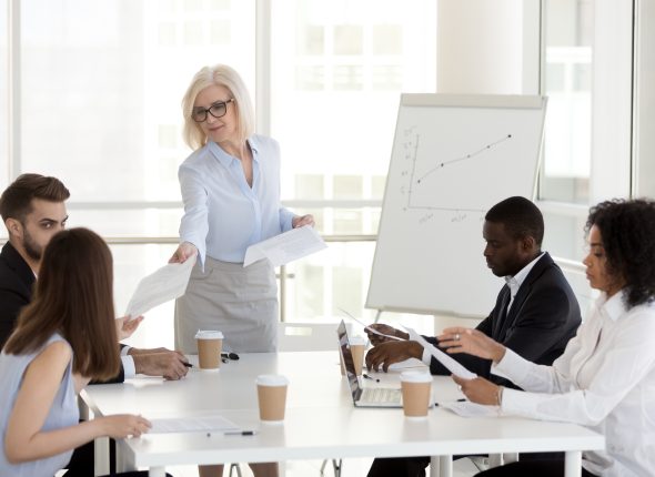 Smiling mature manager giving handouts to diverse employees at meeting